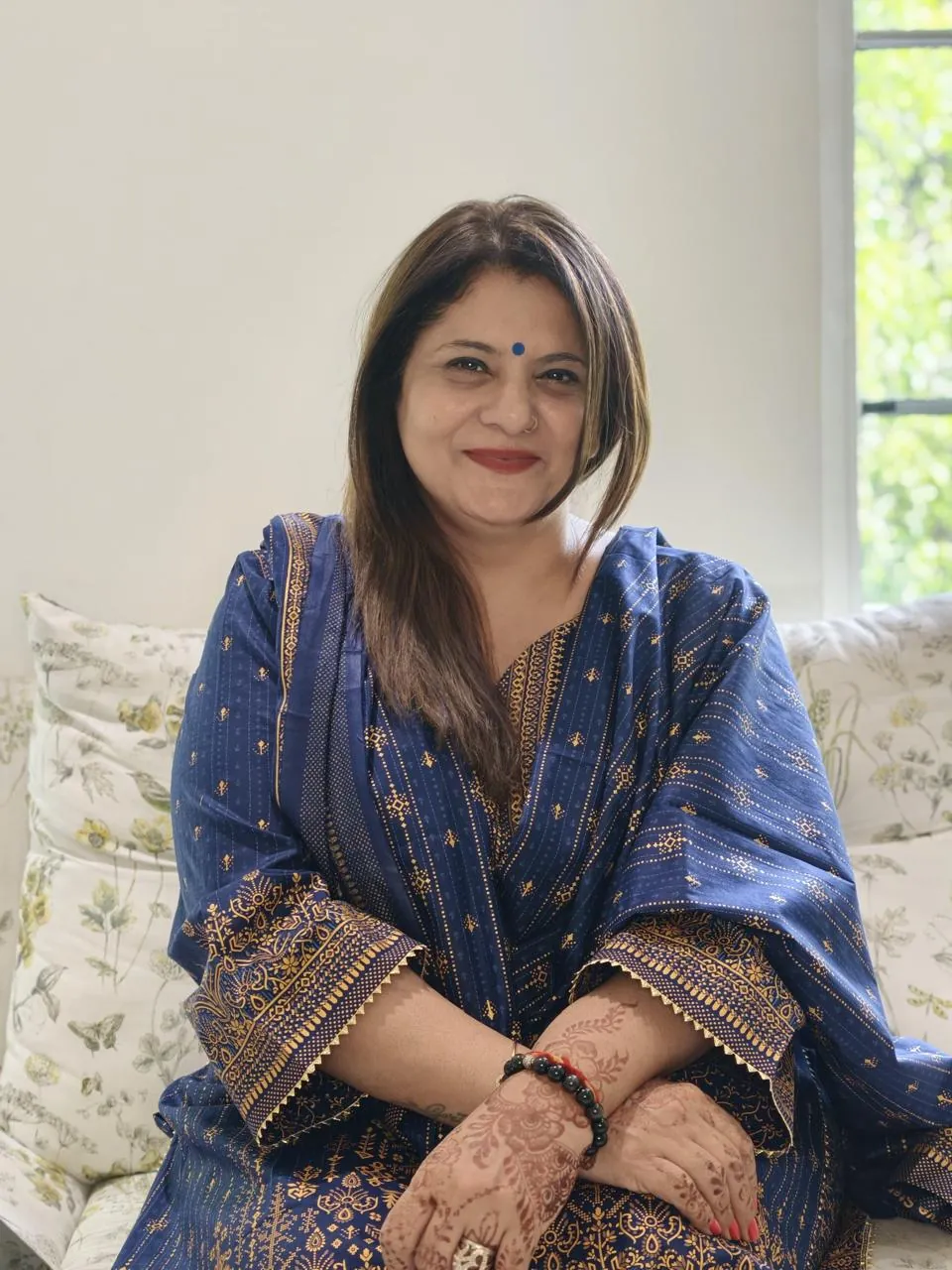 Sapnaa Khemani seated on a floral sofa in a navy kurta with gold block print, henna on her hands