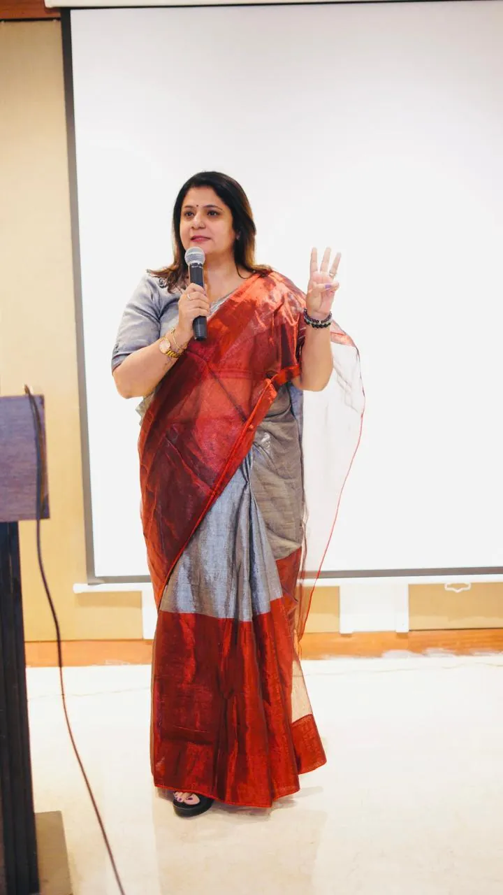 Sapnaa Khemani speaking to an audience with a microphone, a projector screen behind her